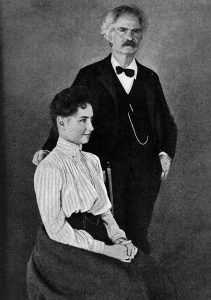 Helen Keller sitting on a chair while Mark Twain stands beside her. The photo is in black and white.
