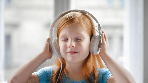 A young girl with red hair is wearing a teal short sleeved shirt and white headphones. Her eyes are closed, and she has a slight smile on her face. She is holding her hands over both earphones and appears to be listening.