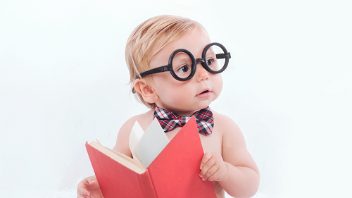 A baby with blond hair is wearing black glasses and a red plaid bowtie. The child is holding a red covered book and looking off to the right.