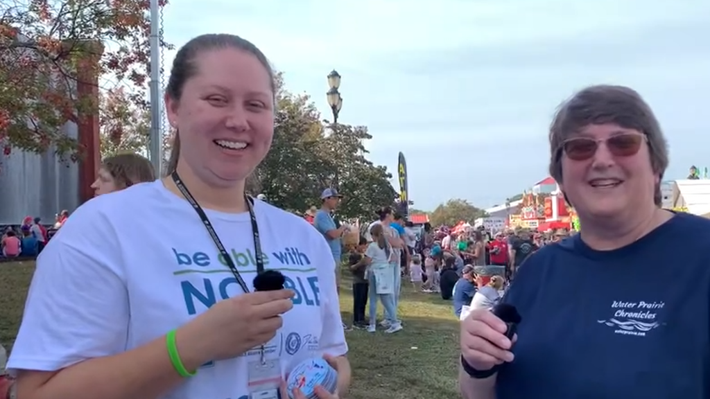 A photo of 2 women standing outside with people in the background walking around the fairgrounds. The woman on the left is white with dark blond hair. She is smiling and wearing a white t-shirt with "be able with NC ABLE" written in blue and green lettering. She is smiling and holding a small black microphone. The woman on the right is white with brown hair and wearing a navy t-shirt with "Water Prairie Chronicles" printed on the left of the chest area. She is wearing sunglasses and is holding a small black microphone.