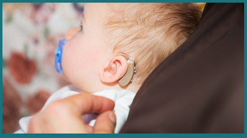 A photo of a baby wearing a hearing aid in their left ear. The side profile of the child is showing, and the baby has a blue pacifier in his mouth. His hair is blond, and he is wearing a white shirt.