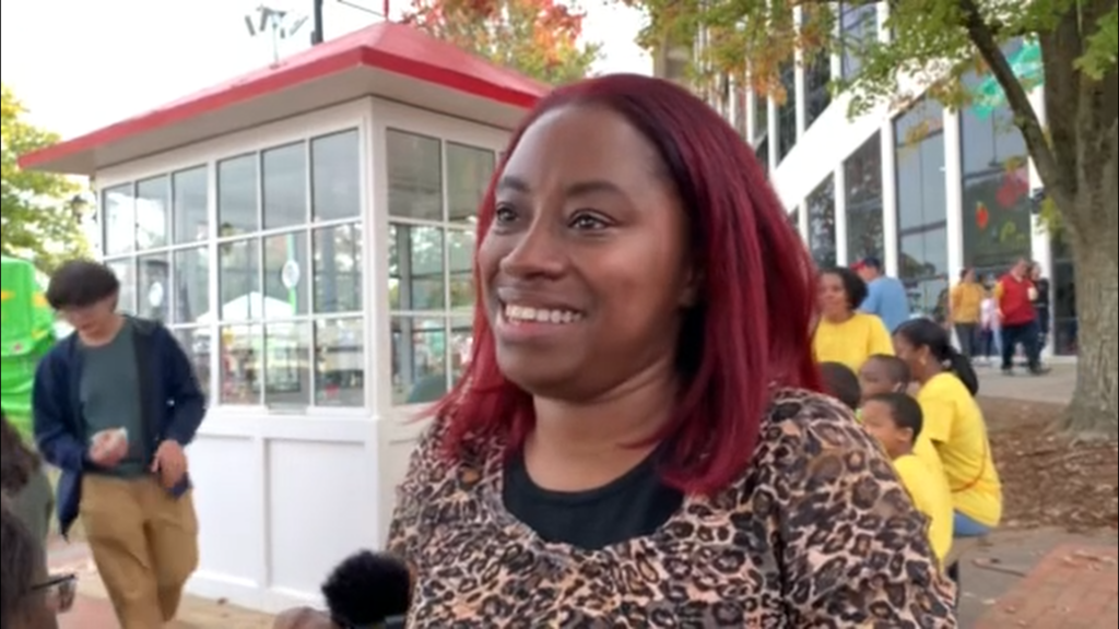 An African-American woman with red hair and wearing a leopard print sweater looks to the left of the photo. She is smiling. Behind her, a group of children and their teacher are sitting at a picnic table, and a young man is walking past her as she talks to her interviewer.