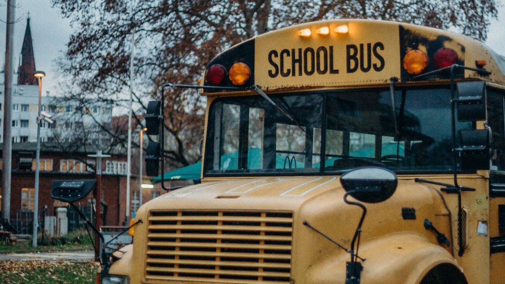 A yellow school bus in front of a building. The bus has lights above the words, "School Bus" printed above the front windshield.
