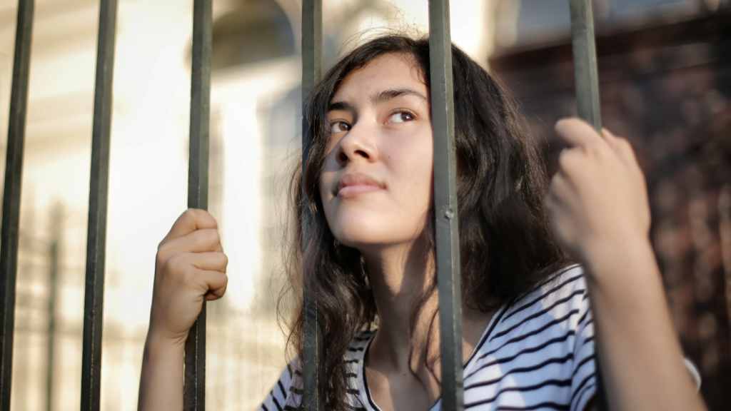 A photo of a girl looking from behind a metal fence. She has brown wavy hair and brown eyes, is looking up and away to the right of the camera, and is wearing a black and white striped shirt.