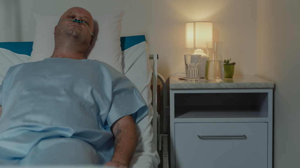 A photo of a man lying in a hospital bed with his head on a white pillow and a nightstand is beside his bed with a small white lamp lit and sitting on the table. The man has a tube in his nose, is bald, is asleep and is wearing a light blue hospital gown.