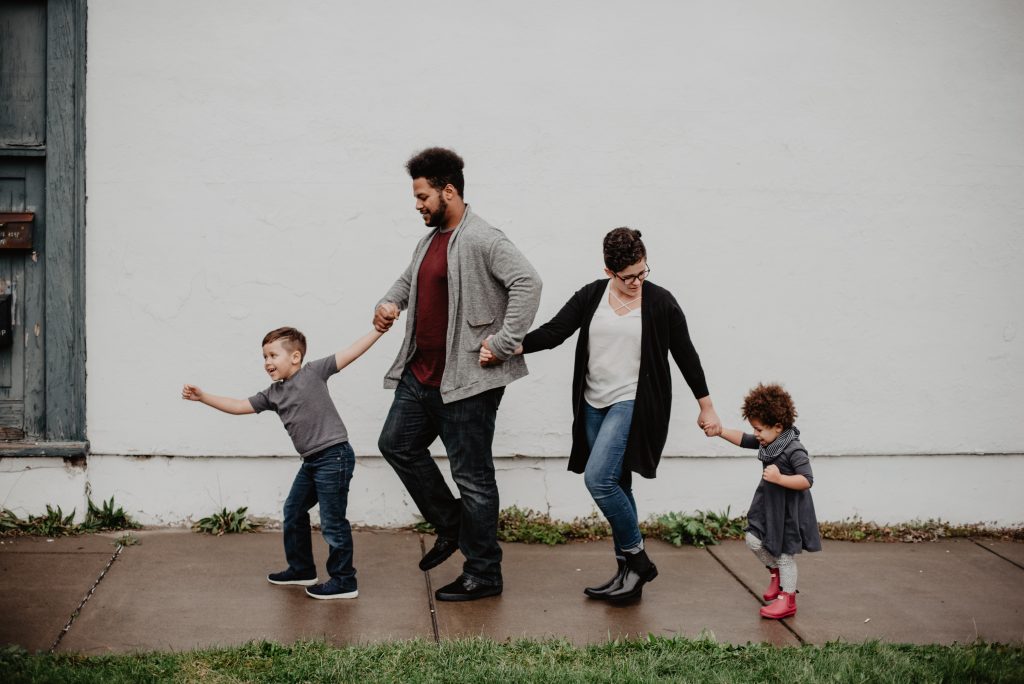 A photo of a family walking on the sidewalk and holding hands. A small boy is in front and is followed by a man with a short black beard who is followed by a woman with short dark hair and holding the hand of a very young girl with short curly dark hair.