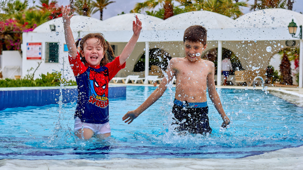 A photo of 2 children playing in a pool. A girl is on the left wearing a Spiderman swim shirt with her arms up above her shoulders as if she has just tossed water into the air. She is smiling with her eyes closed and has shoulder length brown hair. A boy is on the right with short dark hair, wearing dark blue swim shorts and has his hands down so they are touching the water. He looks as if he is about to scoop water into the air.