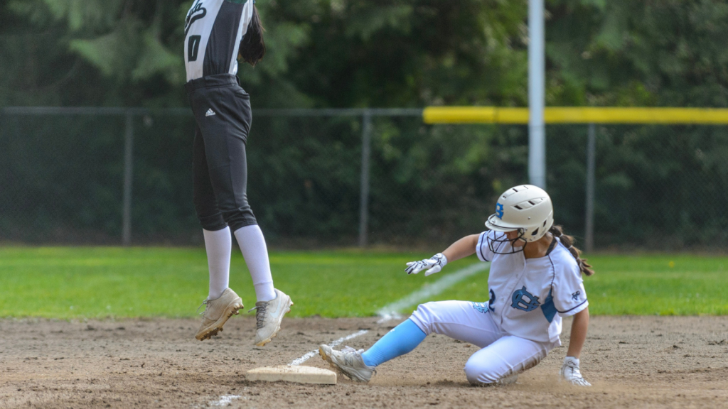 A photo of a softball game with a player sliding into the base. The player is wearing a white uniform and helmet with blue trim. There is another player who is jumping into the air as if to catch a ball. Her torso and legs are in the photo, and she is wering a dark green uniform with white cleats.
