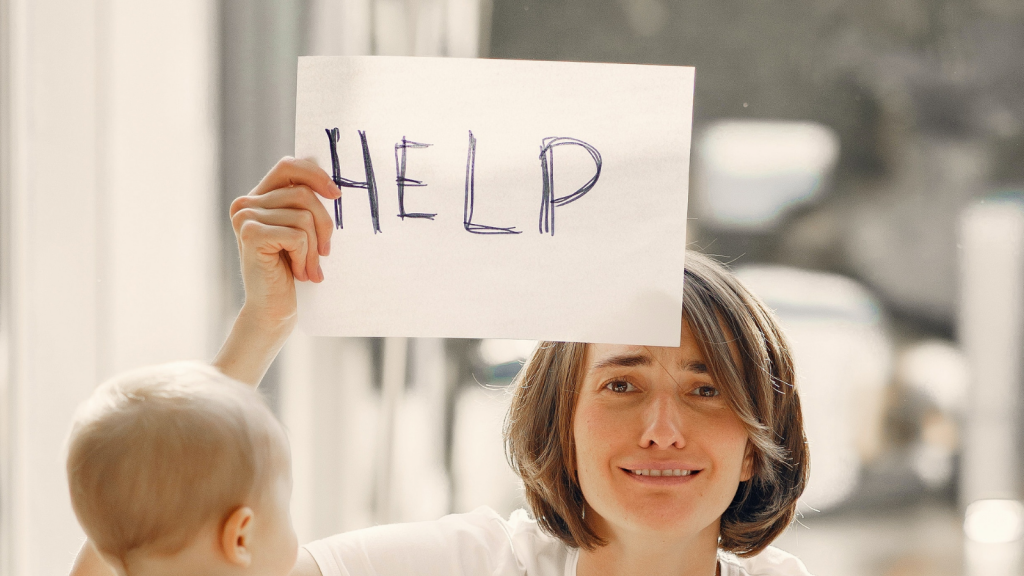 A woman with a stressed look on her face and a child in front of her holds up a white piece of paper with the word "HELP" written on it.