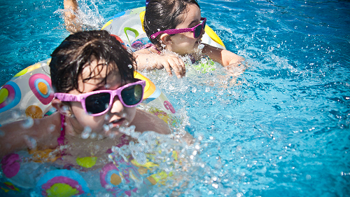 2 girls swimming in a pool with bright blue water. The girls are wearing pink plastic sunglasses and are swimming with pool rings around their waist.