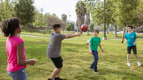 A photo of 4 young teenagers in a park playing with a soccer ball. The teens are standing in a line and appear to be throwing the ball from person to person.