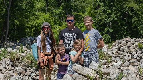 A family of 5 stands to pose for a photo while out hiking. There are rocks in the foreground and green trees in the background. The family are wearing t-shirts and shorts.