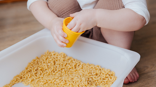 A photo of a young child playing with a yellow cup in a white tub full of dried elbow pasta.