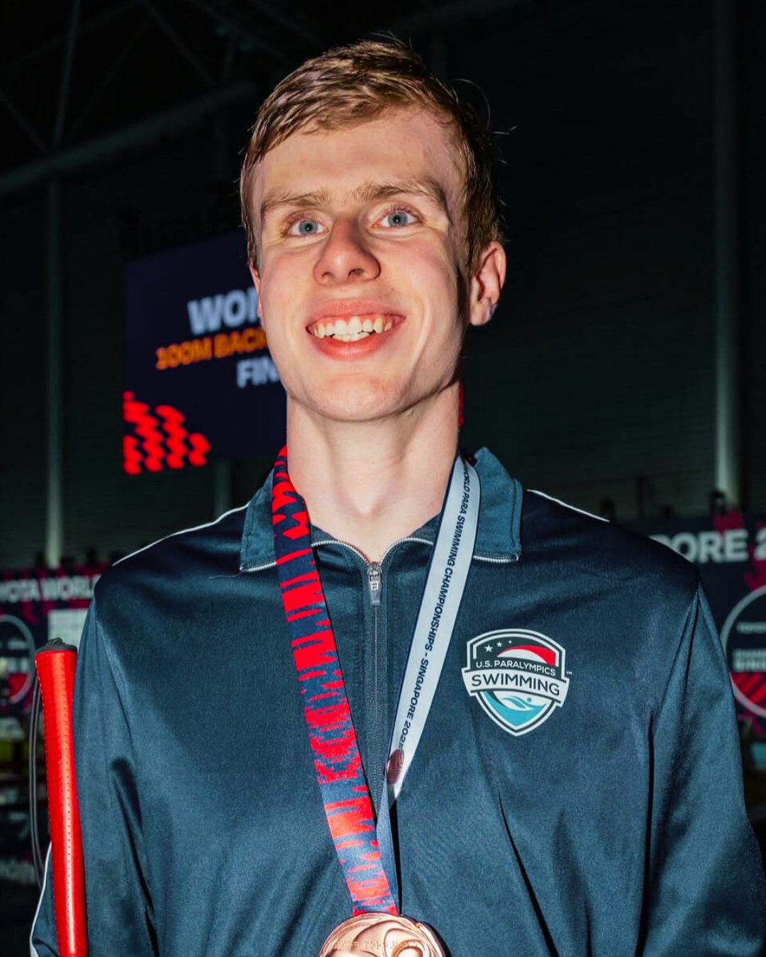 A tall young man with short blond hair wearing a bronze medal around his neck and holding a white cane.