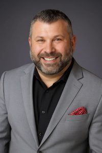 A photo of a man with short dark hair, beard, and mustache is wearing a black shirt with a gray suit coat.